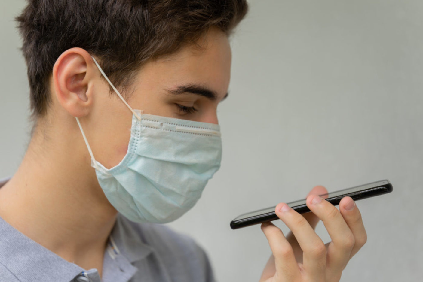 A young man in a disposable medical protective mask on his face is talking on the phone. Teen hold phone speak on speakerphone using virtual digital voice recognition assistant search.