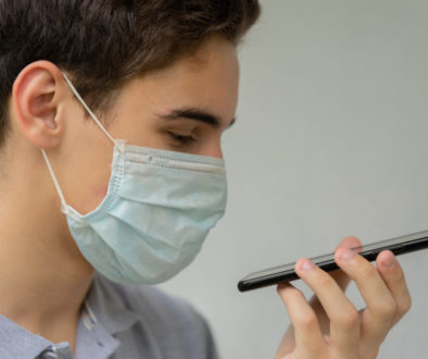 A young man in a disposable medical protective mask on his face is talking on the phone. Teen hold phone speak on speakerphone using virtual digital voice recognition assistant search.