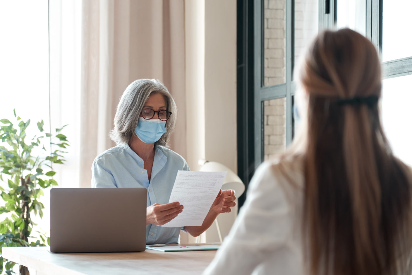 Old hr manager wearing face mask checking female applicant cv at job interview.