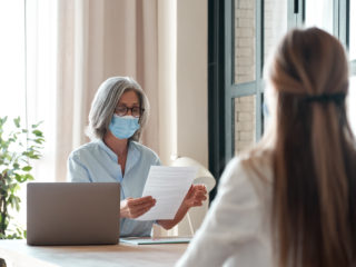 Old hr manager wearing face mask checking female applicant cv at job interview.