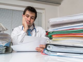 Bureaucracy in medicine concept. Tired overworked doctor is reading medical report. Many documents on desk.