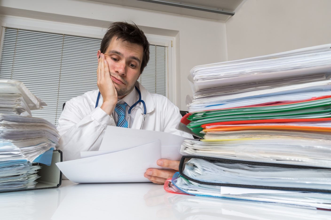 Bureaucracy in medicine concept. Tired overworked doctor is reading medical report. Many documents on desk.